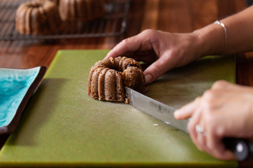 Image of caucasian hands using a knife to cut a pumpkin chocolate chip mini bundt on a green cutting board.